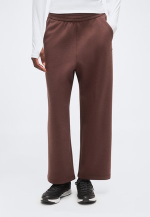 Persona vistiendo pantalones anchos marrones con bolsillos laterales y zapatillas deportivas negras, de pie frente a un fondo liso.