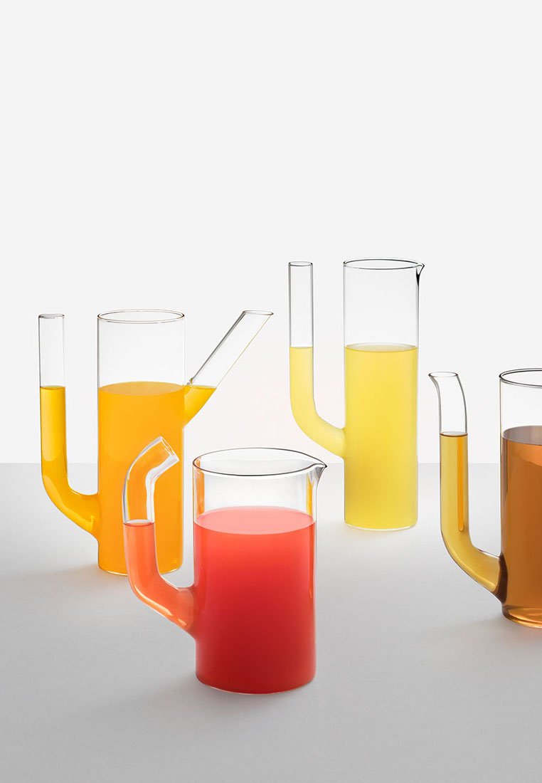 Pięć przezroczystych szklanych dzbanków z unikalnie nachylonymi dziobkami, z których każdy jest wypełniony sokiem o innym kolorze, ustawionych na białej powierzchni.