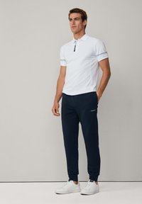Polo bianco con colletto decorativo e strisce blu navy, abbinato a pantaloni jogger blu navy con tasche laterali e logo. Sneakers bianche completano l'outfit.