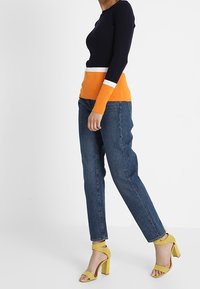 Top de punto con mangas de canalé en azul marino y naranja, con detalles en blanco, combinado con vaqueros de mezclilla azul de tiro alto y sandalias amarillas con tacón cuadrado.
