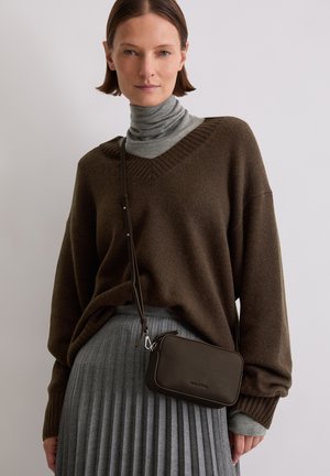 Woman wearing brown oversized sweater over gray turtleneck with gray pleated skirt and small dark brown crossbody bag.