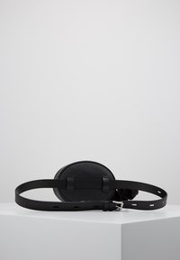 Cirkulär svart läderväska med en slät yta och justerbart bälte. Har två horisontella remmar och ett silverfärgat spänne.