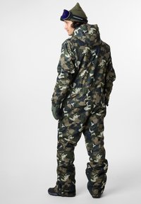 Vihreän sävyissä oleva camouflagen ykköspuku, jossa on kestävä rakenne, hupullinen muotoilu, säädettävät hihansuut ja vetoketjullinen tasku selässä.