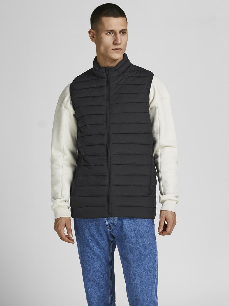 Zwarte gequiltede vest met hoge kraag, ritssluiting aan de voorkant en zijzakken, gedragen over een witte lange mouwenshirt en blauwe jeans. Soepele textuur.