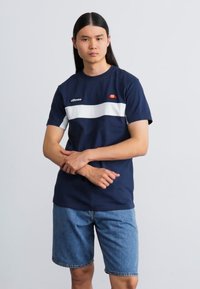 Marineblå kortærmet t-shirt med hvid vandret stribe og logo på brystet, parret med denimshorts, stående mod en neutral baggrund.