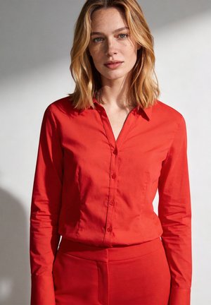 Frau mit schulterlangem blonden Haar, die ein rotes Hemd mit Knöpfen und eine rote Hose trägt und vor einem hellen Hintergrund steht.