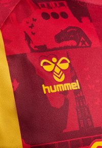 Röd texturerad tyg med gula accenter som har "hummel"-logotypen. Bakgrund innehåller olika mörka silhuetter och mönster.