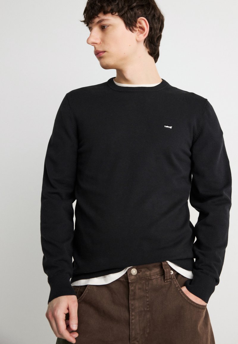 Jeune homme portant un pull noir à manches longues par-dessus une chemise blanche, avec un pantalon marron, regardant sur le côté la main dans la poche.