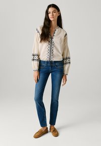 Young woman with long dark hair wearing a cream blouse with blue embroidery, blue jeans, and brown suede moccasin shoes, standing against gray background.