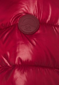 Giubbotto esterno imbottito di colore rosso con texture lucida, design trapuntato e una toppa rotonda in pelle marrone con grafica di montagna e dettagli del marchio.