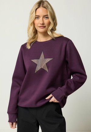 Blond kvinde iført en lilla sweatshirt med en stor leopardprint-stjerne på fronten, poserende med den ene hånd i lommen på sine sorte bukser.
