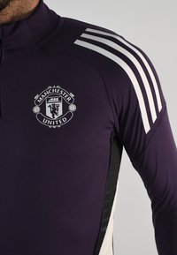 Camiseta deportiva morada con un alto cuello, rayas blancas de Adidas en los hombros y el logo del Manchester United en gris en el frente.