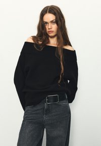 Schwarzer Schulterfreier Pullover mit geripptem Ausschnitt; getragen über hoch taillierter dunkelgrauer Jeans. Schwarzer Gürtel mit quadratischer silberner Schnalle.