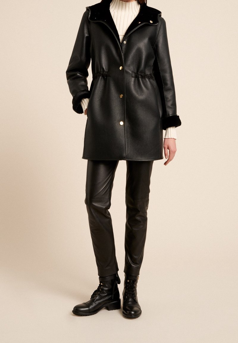 Abrigo de piel sintética negra con botones dorados a presión, cintura elástica y detalles de piel negra en el cuello y los puños, combinado con pantalones de cuero negros y botas.