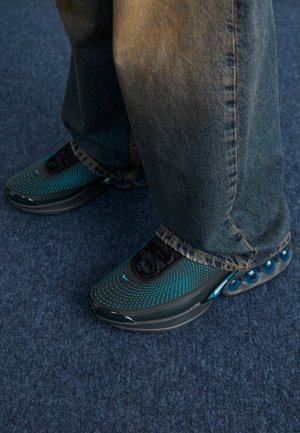 Personne portant un jean ample et des chaussures de sport noires à pois bleus, debout sur un tapis bleu foncé.