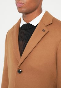 Uomo che indossa un cappotto color cammello sopra un maglione di lana nero e una camicia bianca con colletto, con un singolo bottone nero visibile sul cappotto.