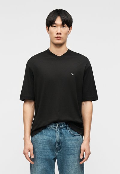 Νεαρός άνδρας όρθιος, φοράει μαύρο T-shirt με V-λαιμόκοψη και μικρό λογότυπο, καθώς και μπλε τζιν, σε απλό φόντο.