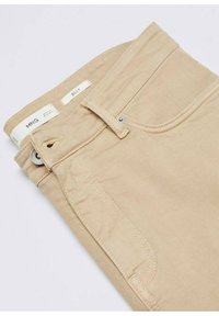 Pantalones de algodón beige con textura suave, que cuentan con un cierre de botón, lazos para cinturón y bolsillos frontales. La etiqueta dice "MNG BILLY."
