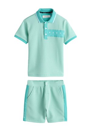 Niño de pie vestido con camisa polo de color verde claro con estampado y pantalones cortos a juego con zapatillas blancas contra un fondo liso.
