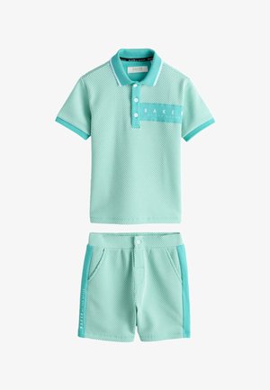 Dreng står iført lysegrøn mønstret polo t-shirt og matchende shorts med hvide sneakers mod ensfarvet baggrund.