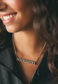 Guldkæde med inskriptionen "ROCK&ROLL" i krystalaccentuerede bogstaver. Glat overflade, sat mod en mørk, silkeagtig top.