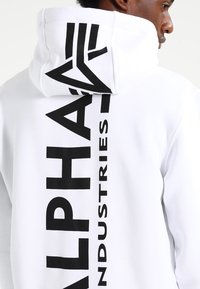 Witte hoodie met een grote zwarte "ALPHA INDUSTRIES" opdruk op de achterkant, voorzien van een trekkoordkap en een gladde, gestructureerde stof.