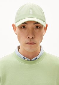 Casquette vert clair avec logo brodé, associée à un sweat-shirt vert clair et une chemise à col bleue en dessous. Gros plan sur le visage.