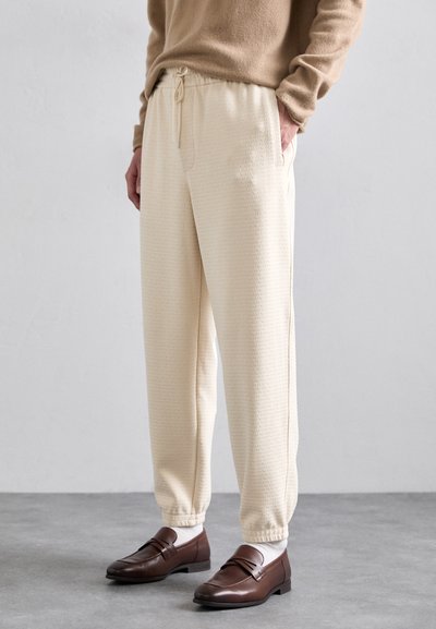 Cream-colored joggers with a textured pattern, featuring an elastic waistband and side pockets, worn with brown loafers and white socks.
