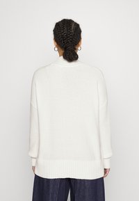 Pull blanc en maille avec un col montant, un ourlet côtelé et des épaules tombantes, présentant une finition texturée et une coupe décontractée.