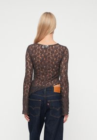 Blusa de encaje marrón de manga larga con un diseño ajustado. Presenta un escote redondeado y una textura transparente, combinada con pantalones de mezclilla oscuros y un parche en la cintura.