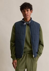 Marineblaue gesteppte Weste mit hohem Kragen und Frontreißverschluss, kombiniert mit einem olivgrünen Pullover und passenden Hosen, ausgestattet mit Taschen und gestrickten Bündchen.