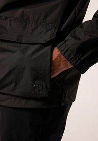Zwarte waterdichte jas met een gladde textuur, uitgerust met een elastische manchette en een zichtbaar logopatch op de zak. Casual ontwerp.