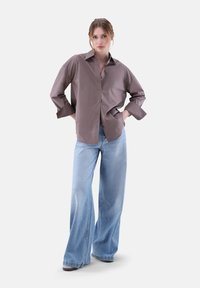 Camicia button-up marrone oversize realizzata in tessuto liscio, abbinata a jeans azzurri a gamba larga; presenta un colletto prominente e una vestibilità ampia.