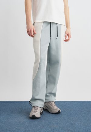 Persona vestida con pantalones bicolor gris claro y blanco con cordones negros y zapatillas Nike grises, de pie sobre una alfombra azul.