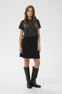 Jauna moteris dėvi blizgantį juodą trumpomis rankovėmis viršų, juodą A formos sijoną ir juodus odinius aulinukus iki kelių, stovinti ant balto fono.