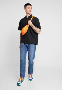 Camisa preta de manga curta com botões, jeans de corte reto azul, bolsa transversal laranja vibrante e ténis multicoloridos com um design dinâmico.