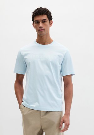 Mann mit lockigem, dunklem Haar, der ein hellblaues kurzärmeliges Hemd und beige Hosen trägt und mit einer Hand in der Tasche steht.