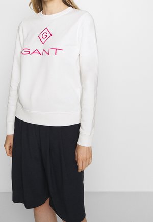 Kvinde iført en hvid sweatshirt med pink GANT-logo og sort knælængde nederdel, stående mod en ensartet lys baggrund.