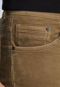 Los pantalones de pana marrones cuentan con un bolsillo trasero con un logotipo de ala bordado y un botón de metal, que exhibe una textura acanalada y un material duradero.
