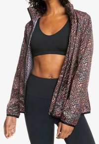 Veste légère avec un motif floral en rose et noir, à fermeture éclair et manches longues. Portée sur un soutien-gorge de sport noir et des leggings.