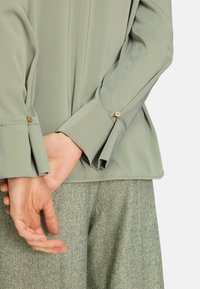 Hellgrüne Bluse mit langen Ärmeln und geknöpften Manschetten, kombiniert mit strukturierten grünen Hosen. Glatte Stoffqualität, lockerer Schnitt und strukturiertes Design.