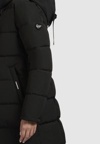 Melna polsterēta jaka ar augstu apkakli, kas aprīkota ar apaļu logo uzlīmi uz piedurknes, kā arī ar regulējamu auklu kapuci un kabatām.