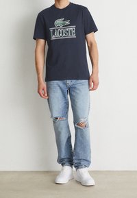 Person wearing a navy Lacoste t-shirt, light blue ripped jeans, and white sneakers, standing against a plain white wall.