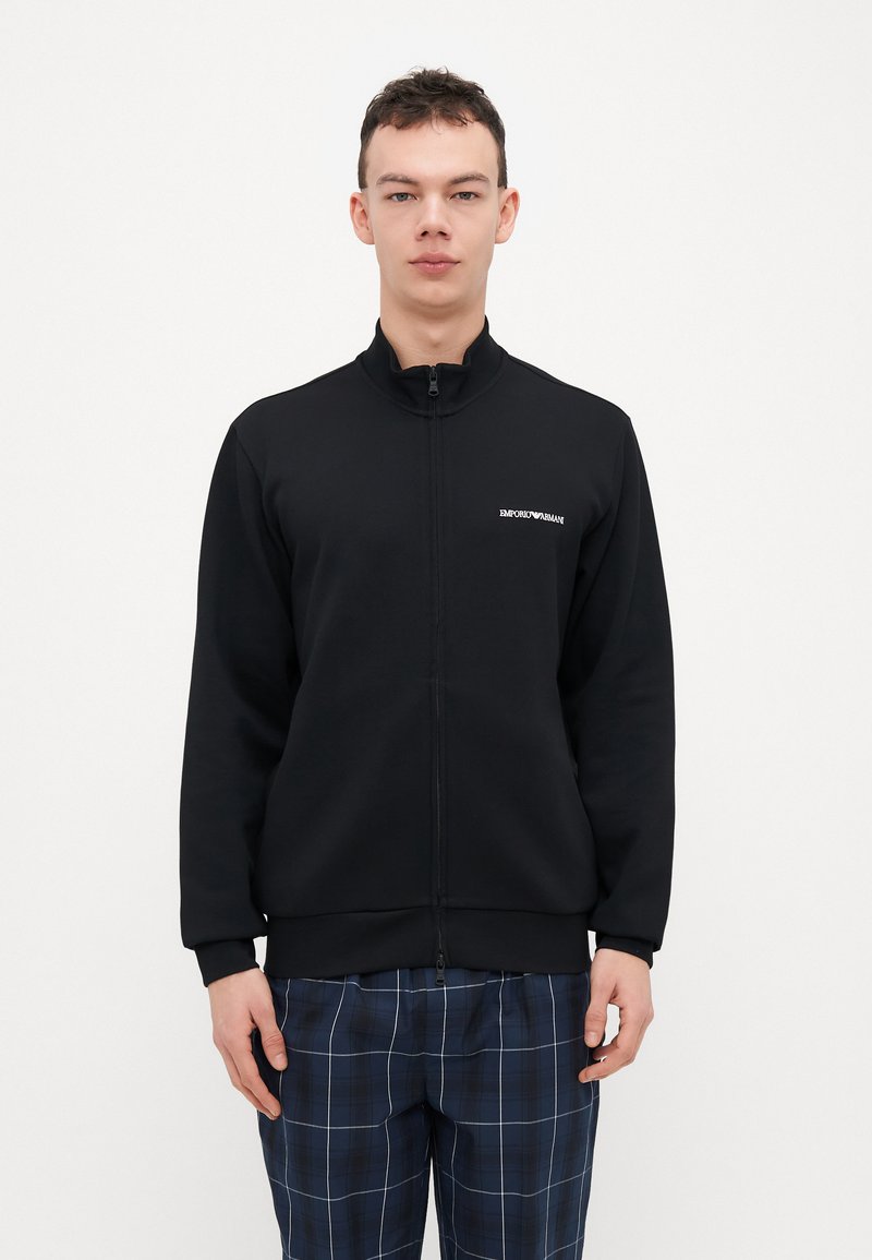 Black zip-up sweatshirt in soft fabric with high collar; features a small white logo on the left chest. Paired with plaid pants.