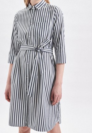 Raidallinen mekko valkoisena ja tummansinisenä, jossa on napitus edessä, kaulus, 3/4-hihat, vyön sidontamahdollisuus ja rintatasku. Sileä kangas.