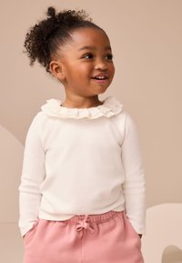 Haut blanc à manches longues avec un col à volants, assorti à un pantalon rose clair à taille ajustable. Le tissu est doux, avec une coupe décontractée.