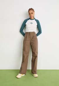 Long-sleeve white and teal top with logo, paired with high-waisted brown trousers. Footwear is light yellow sneakers with accents.