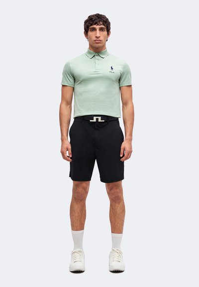 Homme debout face à l’avant, portant un polo vert clair, un short noir avec ceinture, des chaussettes blanches et des baskets blanches sur un fond uni.