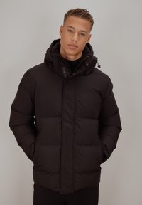 Svart polstret jakke med høy krage, glidelåslukking foran og justerbar hette. Har quiltepartier og sidelommer. Glatt tekstur.