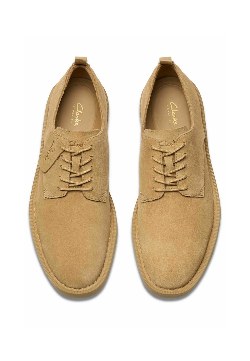 Zapatos de ante beige con punta redonda, que presentan cordones decorativos, una suela texturizada y un logo distintivo en la lengüeta y el costado.